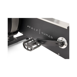 Close-up of a black exercise bike pedal attached to a metal crank arm, with the words "HALF HUMAN" printed on the bike's frame in white letters.
