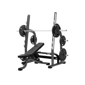 The JORDAN Olympic Adjustable Multi-Bench – a commercial bench press station by Jordan – features an attached barbell rack with loaded weight plates and extra plates stored on side pegs, all shown against a white background.