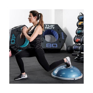 A woman in athletic wear performs a lunge with her back foot on a Jordan BOSU® Balance Trainer (Includes Pump), targeting core development in a gym equipped with stacked weights, exercise mats, and other fitness gear.