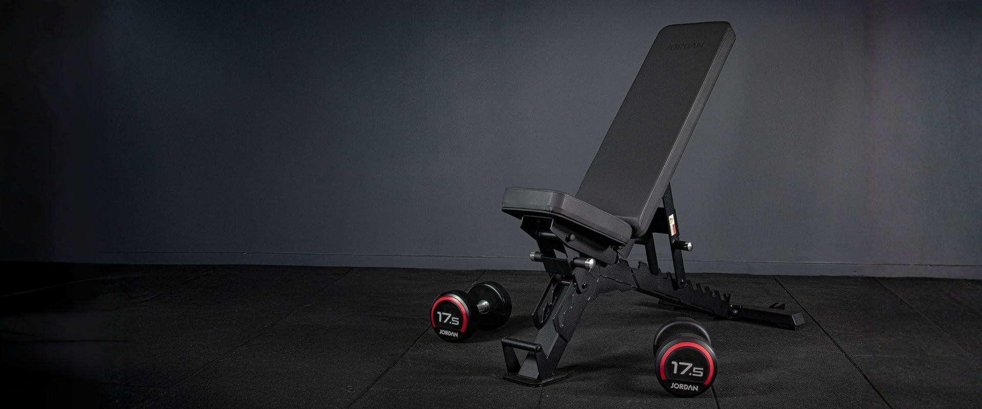 Black adjustable weight bench with red weight plates on a dark floor.