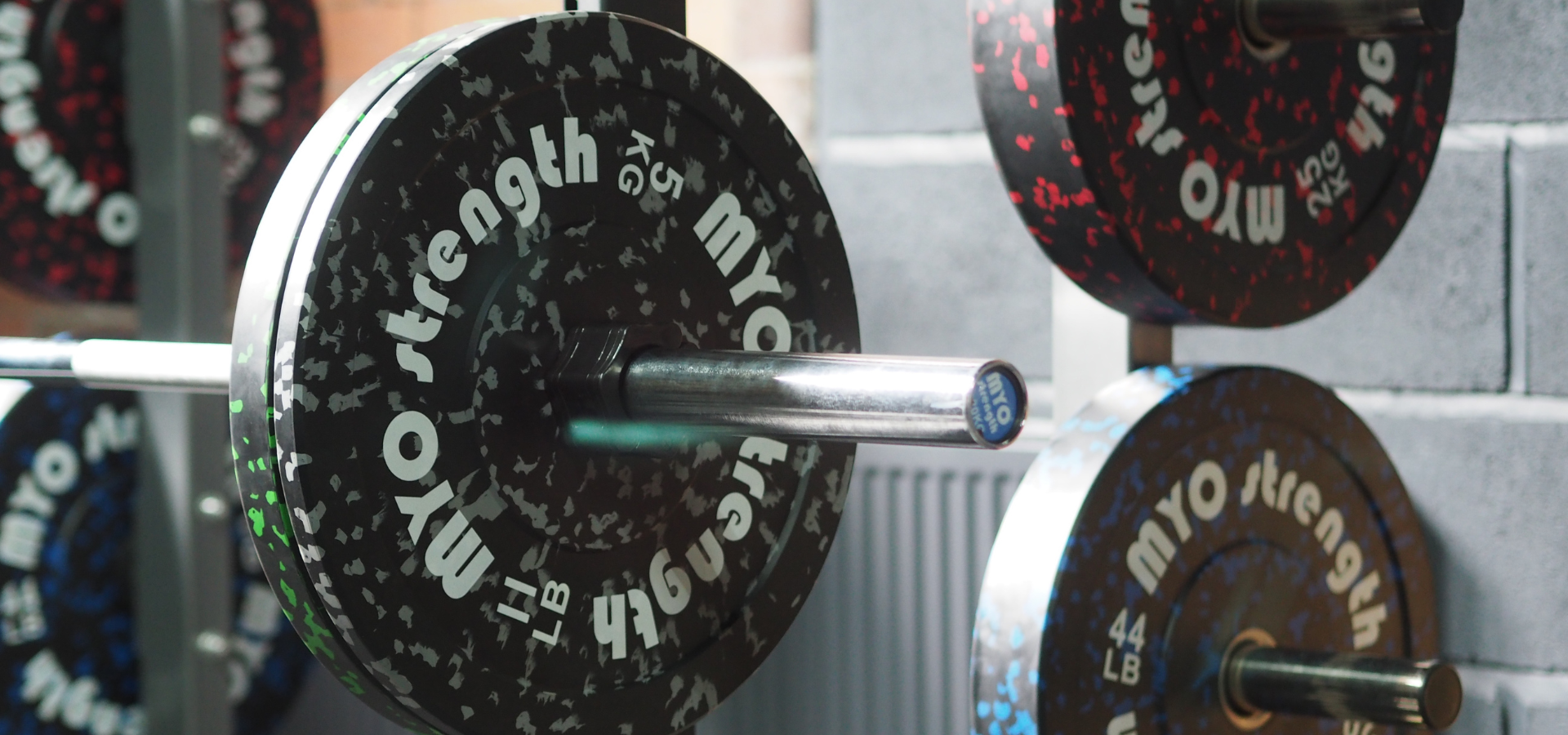 Barbell with weight plates labeled 'MYO Strength' on a gray wall background