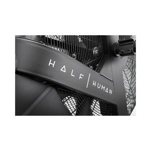 Close-up of a black metal fitness machine with the words "HALF HUMAN" in white, modern lettering on its frame. Mesh and structural details of the machine are partially visible.