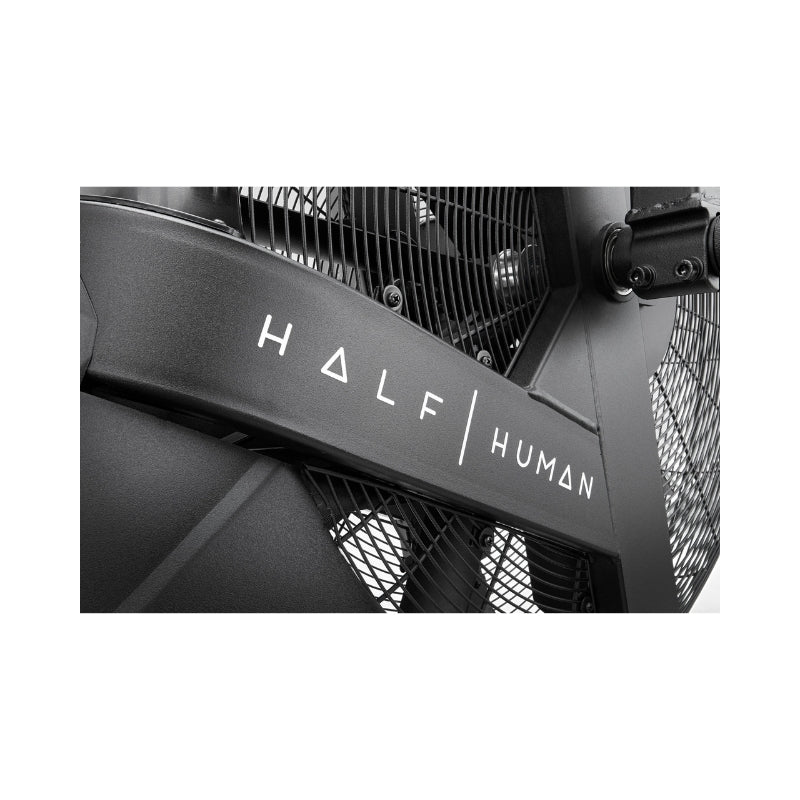 Close-up of a black metal fitness machine with the words "HALF HUMAN" in white, modern lettering on its frame. Mesh and structural details of the machine are partially visible.