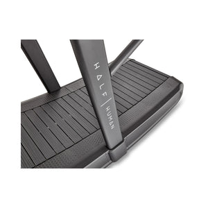 Close-up view of a black treadmill deck with the brand name "HALF HUMAN" printed vertically on the side support bar. The surface of the treadmill features textured, slatted running panels.