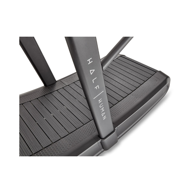Close-up view of a black treadmill deck with the brand name "HALF HUMAN" printed vertically on the side support bar. The surface of the treadmill features textured, slatted running panels.