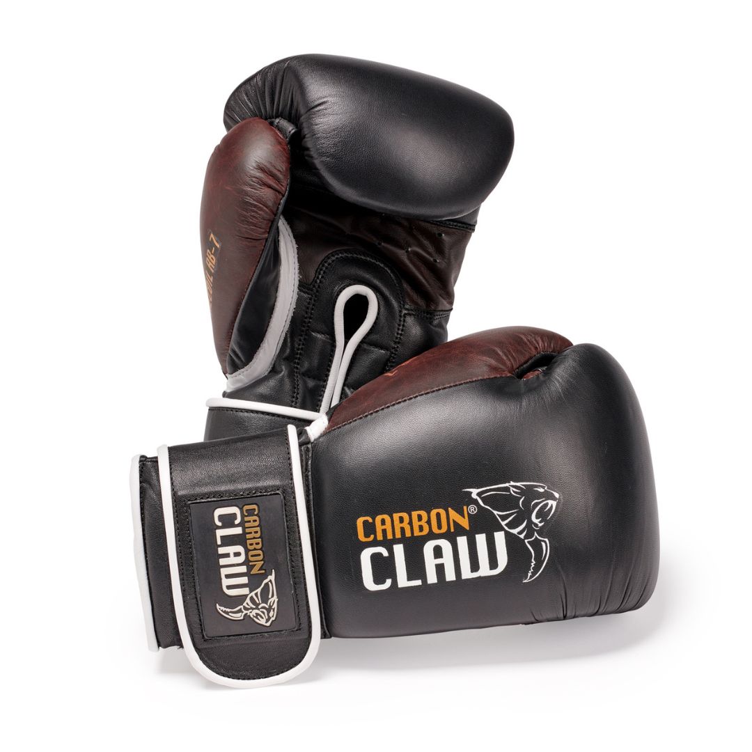 The Carbon Claw Recoil RB-7 leather spar training gloves are sleek in black and maroon, with a white strap for comfort. Expertly designed for wrist support, they feature advanced layered foam technology, one upright and the other resting on its side.